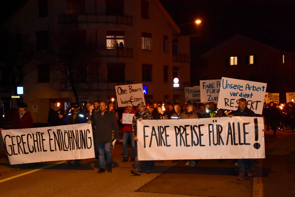 Friedliche Mahnmärsche wie hier in Hinwil am 1. März grenzt Ueli Kuhn klar von Bauernprotesten ab. Man sieht eine Protestkundgebung. Die Menschen tragen Transparente, Kuhglocken und Fackeln.